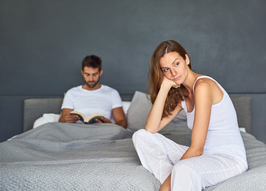 A couple in bed with the woman looking thoughtful and concerned while the man reads a book, depicting a possible scenario of low sex drive or intimacy issues.