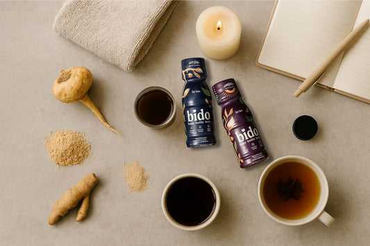 Flat lay of a morning wellness ritual featuring two bido wellness shot bottles, maca root, ashwagandha powder, herbal tea, a towel, a candle, and a journal on a stone surface.